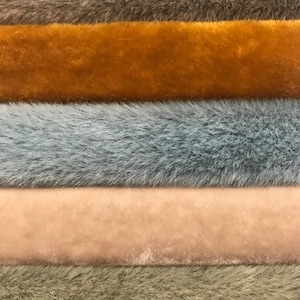 Puede incluir: Primer plano de una colección de telas de piel sintética en varios colores. Los colores incluyen gris, amarillo mostaza, azul-gris, rosa claro y verde oliva. Las telas tienen un aspecto suave y texturizado.