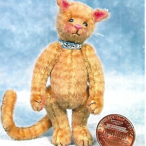 Puede incluir: Un gato de peluche naranja hecho a mano con un collar azul, de pie. El gato tiene detalles rosas en las orejas y la nariz. Una moneda está al lado del gato para la escala. La cola del gato está curvada.