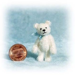 May include: A small, white teddy bear with a blue ribbon around its neck. The bear is standing on a blue surface and is about the size of a UK penny.