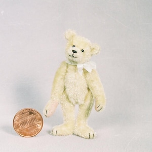 Peut inclure: Un petit ours en peluche crème avec un nœud papillon blanc est debout. L'ours a des yeux noirs et un nez cousu noir. Une pièce de monnaie est à côté de l'ours pour la comparaison de taille. Les pattes de l'ours sont roses.