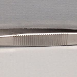 May include: A stainless steel tweezer with a ridged grip and finely serrated tips. The tweezer is designed for precision work, with a flat, rectangular end opposite the tips. The tool is shown against a white and gray background.
