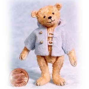 May include: A small, light brown teddy bear wearing a light blue hooded jacket with gold buttons. The bear is standing on a white surface next to a penny.
