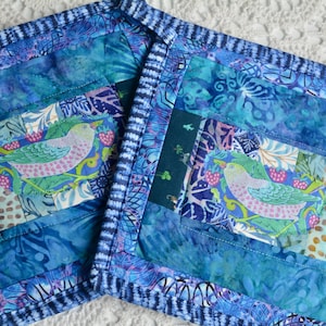May include: Two square quilted fabric potholders featuring a patchwork design in shades of blue, teal, and purple. Each potholder displays a central graphic of a light green bird perched on a branch with pink berries, surrounded by patterned borders.