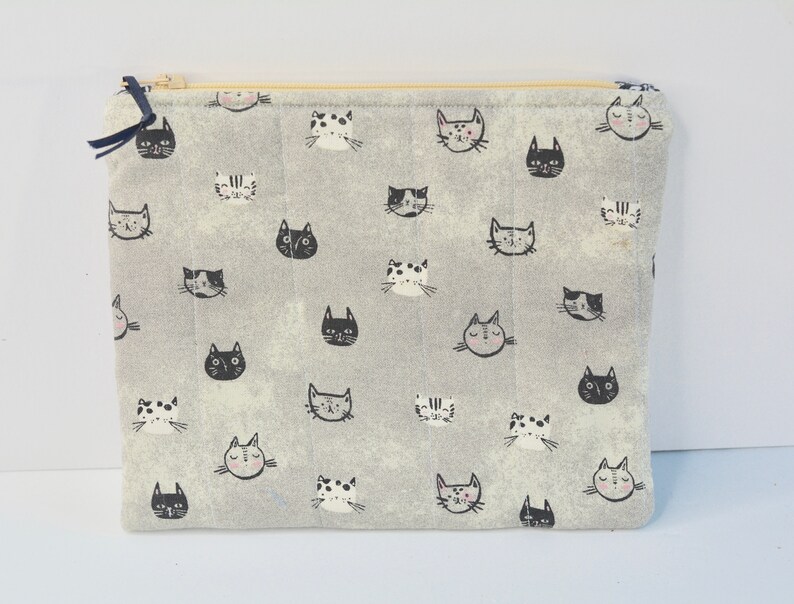 May include: A grey zippered pouch with a repeating pattern of black and white cat faces. The pouch is made of a soft, quilted fabric.