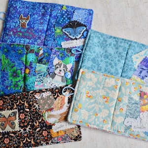 May include: A collection of handmade fabric pouches in various blue floral and patterned designs. Each pouch features unique appliqued details, including butterflies, a fox, and a dog. Some pouches have a loop and button closure.