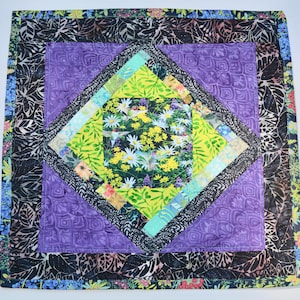 May include: A purple and black quilted square with a floral pattern in the center. The floral pattern is surrounded by a diamond-shaped frame.