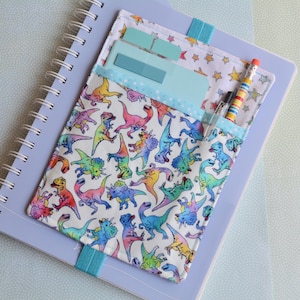 May include: A notebook with a colorful dinosaur-themed cover and a matching pocket. The pocket holds sticky notes, a pen, and a pencil. The notebook is light purple with a spiral binding and a blue elastic band.