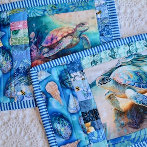 May include: Two square mug rugs with a blue and white striped border. Each rug features a colorful sea turtle design. The background of the rugs includes seashell and ocean-themed patterns. The tag reads "Mug Rug".