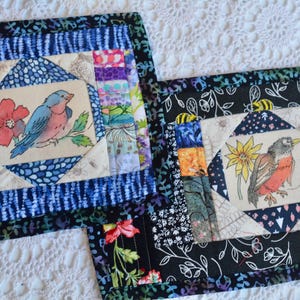 May include: Two square quilted placemats. One features a blue bird and red flower, the other a bird with a yellow flower. Both have a central square with a bird illustration, surrounded by patterned fabric borders.