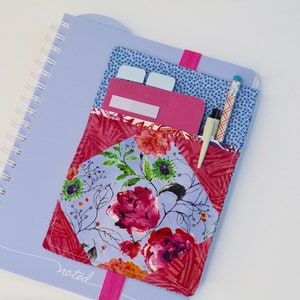 May include: A blue notebook with a floral fabric pocket attached. The pocket has a pen and a few sticky notes inside. The notebook has the word "noted" written on the front.