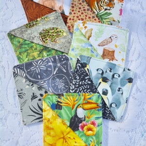 May include: Nine fabric coasters with different animal and nature designs. The coasters are made of a soft, quilted fabric and feature a variety of colors, including green, blue, brown, and yellow. The designs include a frog, a tiger, an owl, a toucan, a fox, a starfish, and penguins.