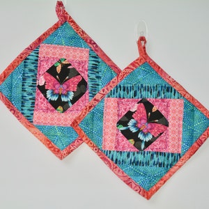 May include: Two quilted hot pads with a pink and blue butterfly design. The hot pads are made of fabric with a patchwork pattern and have a coral-colored border.
