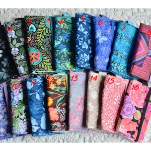 May include: A collection of sixteen fabric-covered notebooks, each with a unique patterned design. The notebooks feature a variety of colors, including blue, green, pink, and purple, with floral, geometric, and abstract patterns. Each notebook has a coordinating elastic band closure.