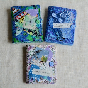 May include: Three handmade fabric needle cases with button closures. The cases feature patchwork designs with butterfly, dragonfly, and bird appliqués. Each case has a unique floral and patterned fabric in shades of blue, purple, and green. The text "Needles & Pins" is on two cases.