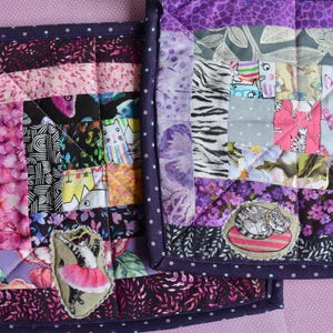 May include: Two handmade quilted pot holders. The pot holders feature a patchwork design with various floral, animal, and geometric patterns in shades of purple, pink, and black. Each pot holder has a dark purple polka dot border.