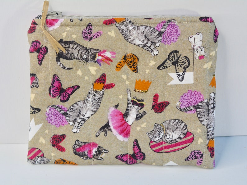May include: A beige zippered pouch with a quilted texture. The pouch features a repeating pattern of cats, butterflies, and flowers in shades of pink, grey, and white.