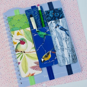 May include: Three fabric pencil holders with elastic bands. The holders feature different bird designs: a hummingbird, a blue jay, and a yellow bird. The holders are attached to a blue notebook.