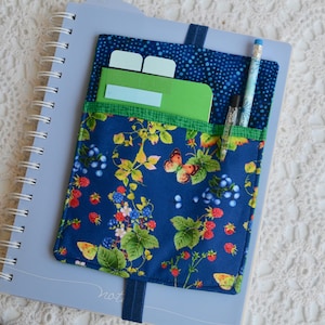 May include: A blue notebook with a fabric pocket featuring a floral design of butterflies, berries, and leaves. The pocket holds a green notebook, two index cards, and pens. A blue pen and a black pen are visible.