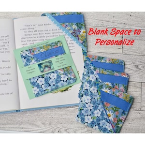 May include: Four blue and white floral fabric corner bookmarks with a blank space to personalize. The bookmark on the left has the text "Corner Bookmark" and "Olivia" written on it. The text "Just One More Chapter" and "Slip onto page corner. Save your place." are also visible on the bookmark.