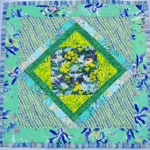 May include: A green and blue quilt with a diamond pattern. The center of the quilt features a floral design with hummingbirds. The quilt is edged with a blue and white trim.