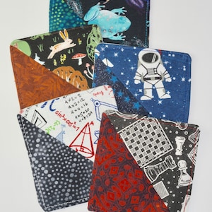 May include: Five fabric squares with different patterns. The squares are sewn together to create a larger square. The patterns include a frog, a rabbit, an astronaut, math equations, and a chessboard.