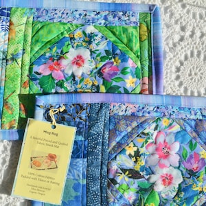 May include: Two quilted mug rugs with floral and patchwork designs. The rugs feature a mix of blue, green, and floral patterns. A yellow tag is attached to one, with text that reads "Mug Rug". The rugs are approximately 8 inches square.