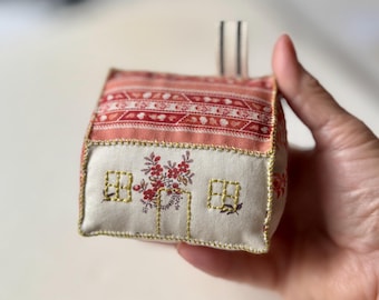 May include: A handmade fabric house with a red and white patterned roof and a cream-colored front. The front features embroidered windows and a floral design above the door. The edges are trimmed with gold stitching, and a striped ribbon is attached to the top.