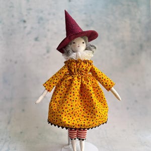 May include: A fabric doll wearing a pointed burgundy hat with a black pattern, a white ruffled collar, and a yellow dress featuring a small black and red floral print. The doll has gray hair, red and white striped leggings, and stands on a white base.