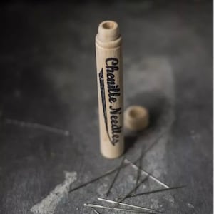 May include: A wooden tube with the text "Chenille Needles" printed on it. The tube is open and several chenille needles are scattered on a gray surface.