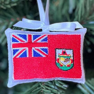 May include: Embroidered Christmas ornament featuring the flag of Bermuda. The ornament is rectangular with a red background and the Union Jack in the upper left corner. The Bermuda coat of arms is on the right. A white ribbon is attached for hanging.