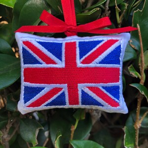 May include: Embroidered Union Jack pillow ornament with red ribbon for hanging. The pillow features a white border and a detailed design of the British flag in red, white, and blue. Perfect for patriotic decor.