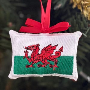 May include: A rectangular fabric ornament featuring the Welsh flag with a white top half and green bottom half, centered with a red dragon embroidery. The ornament is topped with a bright red satin ribbon tied into a bow for hanging purposes.
