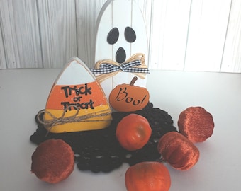 May include: A Halloween tiered tray decor set featuring a white wooden ghost with a black and white gingham bow, a yellow and orange candy corn cutout with the text Trick or Treat, and a small orange pumpkin cutout with the text Boo.