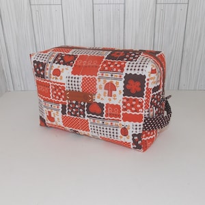 May include: A rectangular red, brown, and white patchwork fabric zippered pouch with a brown leather patch and a brown and white polka dot handle.