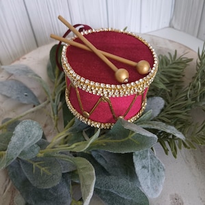 May include: A small, cylindrical drum ornament covered in deep red velvet fabric. The top and bottom edges are trimmed with sparkling clear rhinestones. Gold cord creates a zigzag pattern around the sides. Two thin, light-colored drumsticks rest on top.