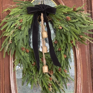 May include: A green pine wreath with a black velvet bow and gold bells hanging from a jute rope. The wreath is decorated with pine cones and is hanging on a wooden door.