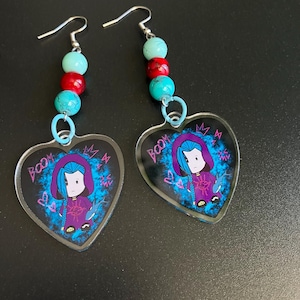 Pode incluir: Um par de brincos em forma de coração com um personagem de desenho animado com cabelo azul e uma roupa roxa. Os brincos têm um design de contas com contas turquesas, vermelhas e turquesas. A palavra "BOOM" é visível.