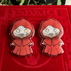 May include: Two red enamel pins depicting a hooded figure with a white face and large eyes. The pins are set against a red background. The figure is wearing a red robe with a hood.