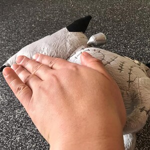 May include: A grey plush toy, possibly a wolf or dog, with black details is shown. The toy has stitched details and a small, silver-coloured element. The image focuses on the toy's texture and design.