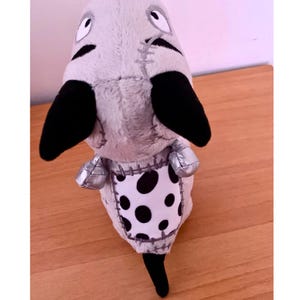 May include: A soft toy creature with a grey body, black ears, and a black tail. It features a white and black polka-dot patch on its belly and silver-coloured feet.