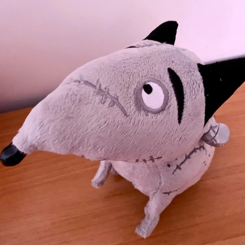 May include: A grey plush dog toy with black ears and nose. The toy features stitched details and a metal-coloured bolt on its neck. The dog has a cartoon design with a large eye and is made of soft material.