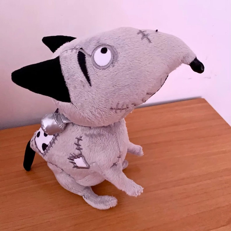 May include: A grey plush dog toy with black ears and a black nose. The toy has stitched patches and a silver collar. The dog has a large, expressive eye and is sitting on a wooden surface.