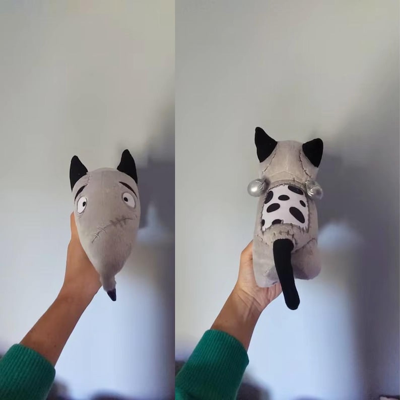 May include: A handmade grey plush toy with black ears and a black tail. The front view shows a sad face, while the back view reveals a spotted patch and silver accents. The toy is held up against a light grey wall.