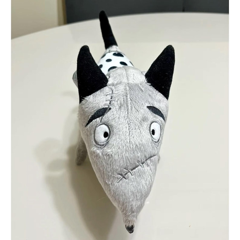 May include: A grey, black, and white plush dog toy. The toy features stitched details, large eyes, and black ears. The tail is black with white spots. The dog has a sad expression.
