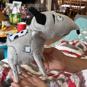 May include: A grey plush dog toy with black ears and a black nose. The toy features a white patch with black spots and a stitched-on patch. The toy is held in a person's hands.