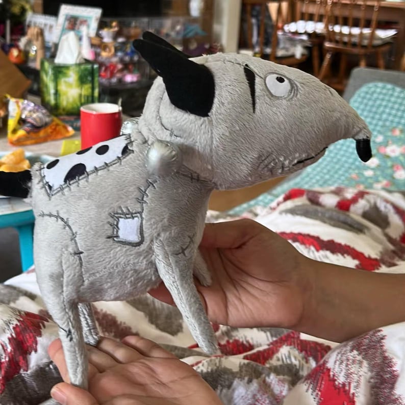 May include: A grey plush dog toy with black ears and a black nose. The toy features a white patch with black spots and a stitched-on patch. The toy is held in a person's hands.