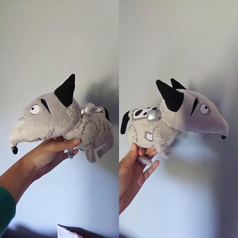 May include: A soft toy dog-like creature in grey with black ears and a long snout. The toy has stitched details and patches, with a small silver accent on its back. The eyes are large and round, giving it a whimsical appearance.