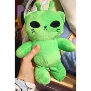 Op de afbeelding: Een groene pluche speelgoed in de vorm van een alien-kat. Het speelgoed heeft grote zwarte ovale ogen, kleine zwarte snorharen en twee kleine antennes. Het pluche is zacht en knuffelig, ideaal voor kinderen of verzamelaars.