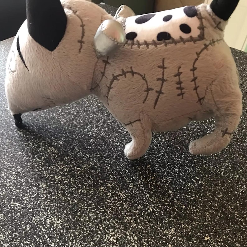 May include: A grey dog-shaped plush toy with black ears and a white and black spotted patch on its back. The toy features stitched details and a silver metallic accent.