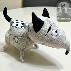 May include: A soft toy dog with a Frankenstein theme. The dog is mainly grey with black ears and tail, and a white and black spotted patch on its back. It features stitched details and a silver accessory. The image includes the text "Frankenweenie".